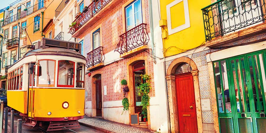 A bright yellow tram travels down the track of a Lisbon street with brightly coloured doors. 